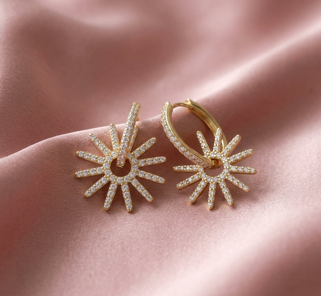 A pair of gold hoop earrings with sunburst-shaped dangling charms paved with small clear stones, displayed on a draped pink satin fabric.