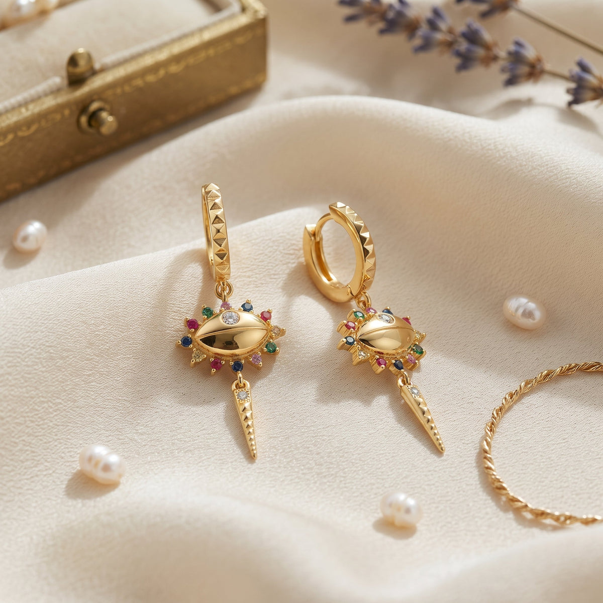 A pair of gold huggie hoop earrings with dangling lightning-bolt charms embellished with colorful gemstones, displayed on a cream fabric surface with decorative pearls, a gold box, and dried lavender.