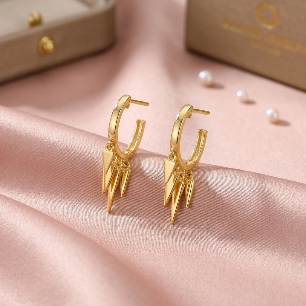 A pair of gold hoop earrings with three pointed triangular dangles, displayed on draped pink satin fabric with pearl-like beads and jewelry boxes in the background.