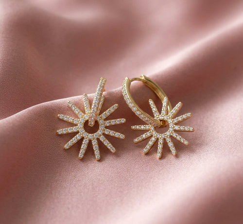 A pair of gold hoop earrings with sunburst-shaped dangling charms paved with small clear stones, displayed on a draped pink satin fabric.
