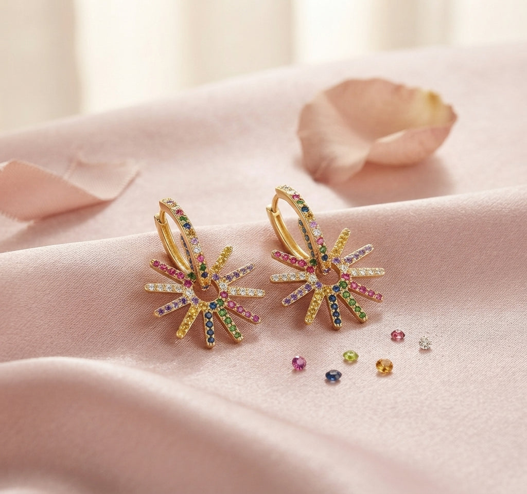 A pair of gold hoop earrings with multi-coloured gemstone sunburst designs, displayed on soft pink satin fabric with a single rose petal and loose accent stones.