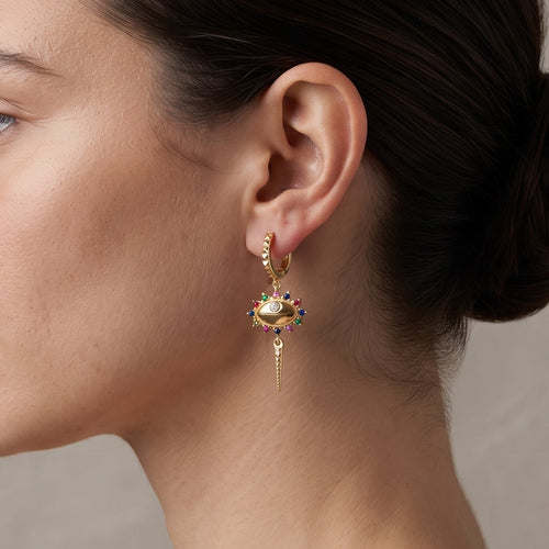 A woman in profile wearing a delicate gold huggie hoop earring with a dangling cross-shaped charm adorned with small colorful gemstones, set against a neutral textured gray background.
