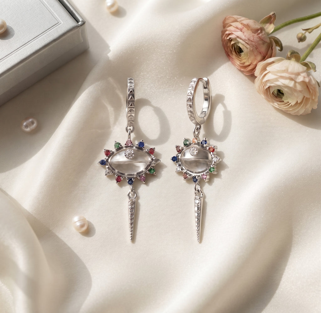 A pair of silver dangle earrings with clear oval center stones and colorful gemstone accents, displayed on draped cream satin fabric with flowers and a small gift box nearby.