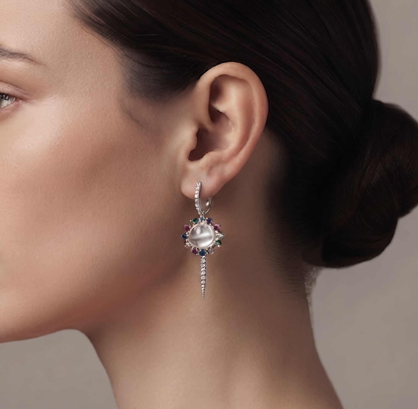A close-up side profile of a woman with dark hair in a bun, wearing a delicate silver-toned dangle earring with a clear central stone and small blue gemstone accents, against a soft gray background.