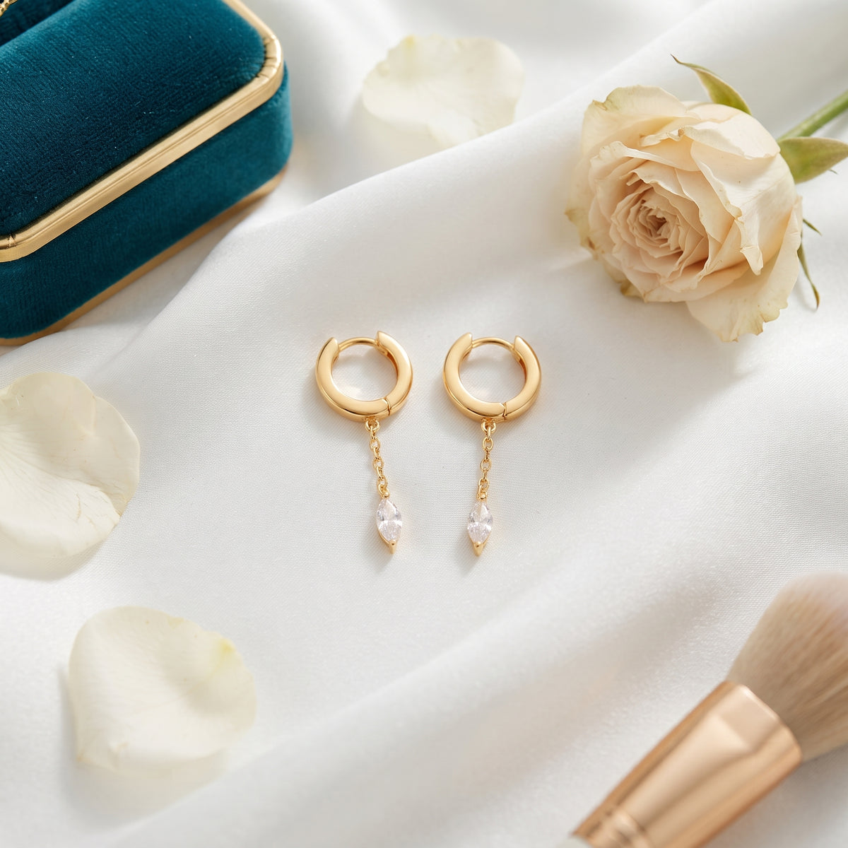A pair of gold huggie hoop earrings with dangling clear marquise-cut crystals, styled on a white satin fabric with rose petals, a cream rose, a teal velvet box, and a makeup brush.