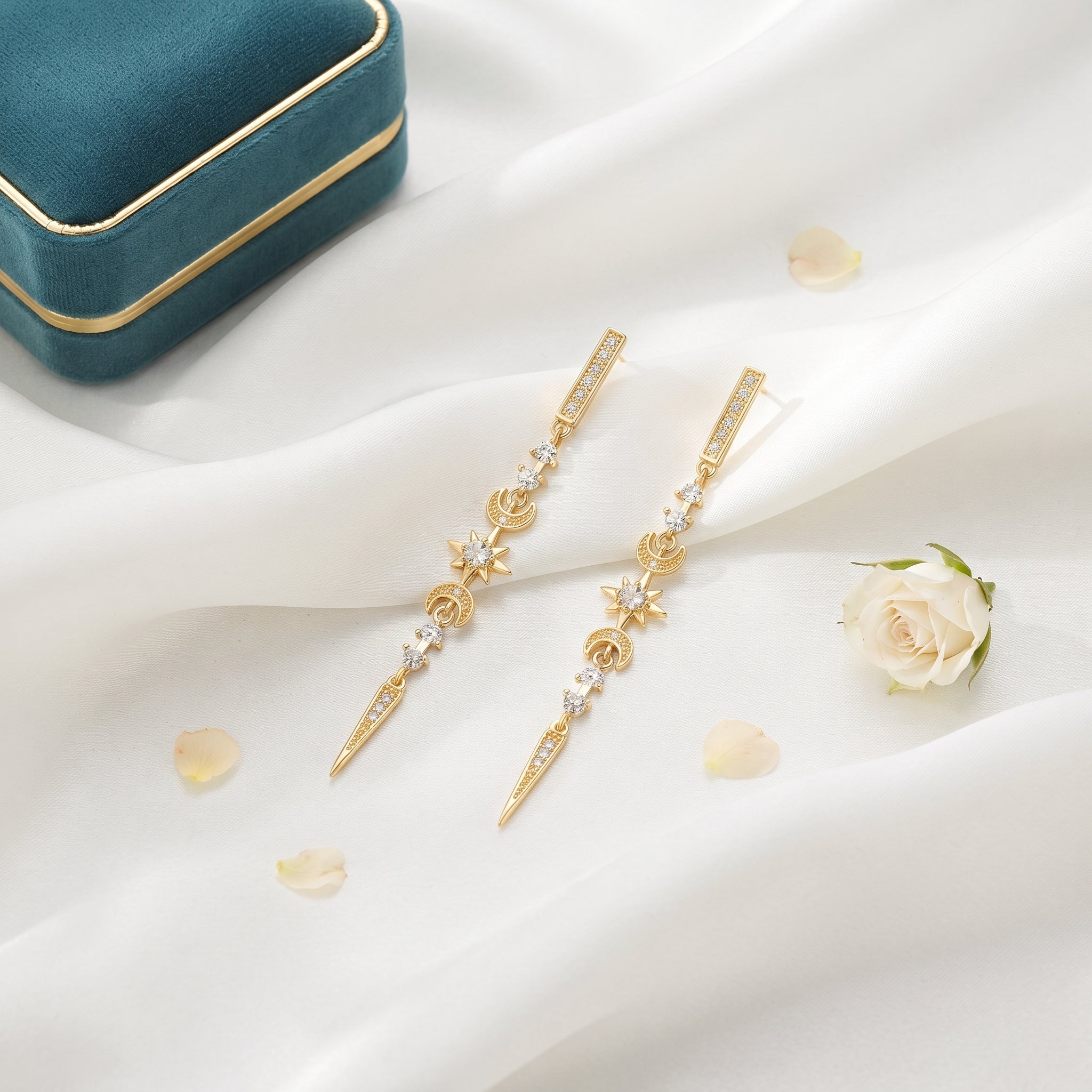 A pair of gold-toned statement earrings with clear gemstones and floral motifs, displayed on draped white fabric with a small white rose and a teal jewelry box in the background.
