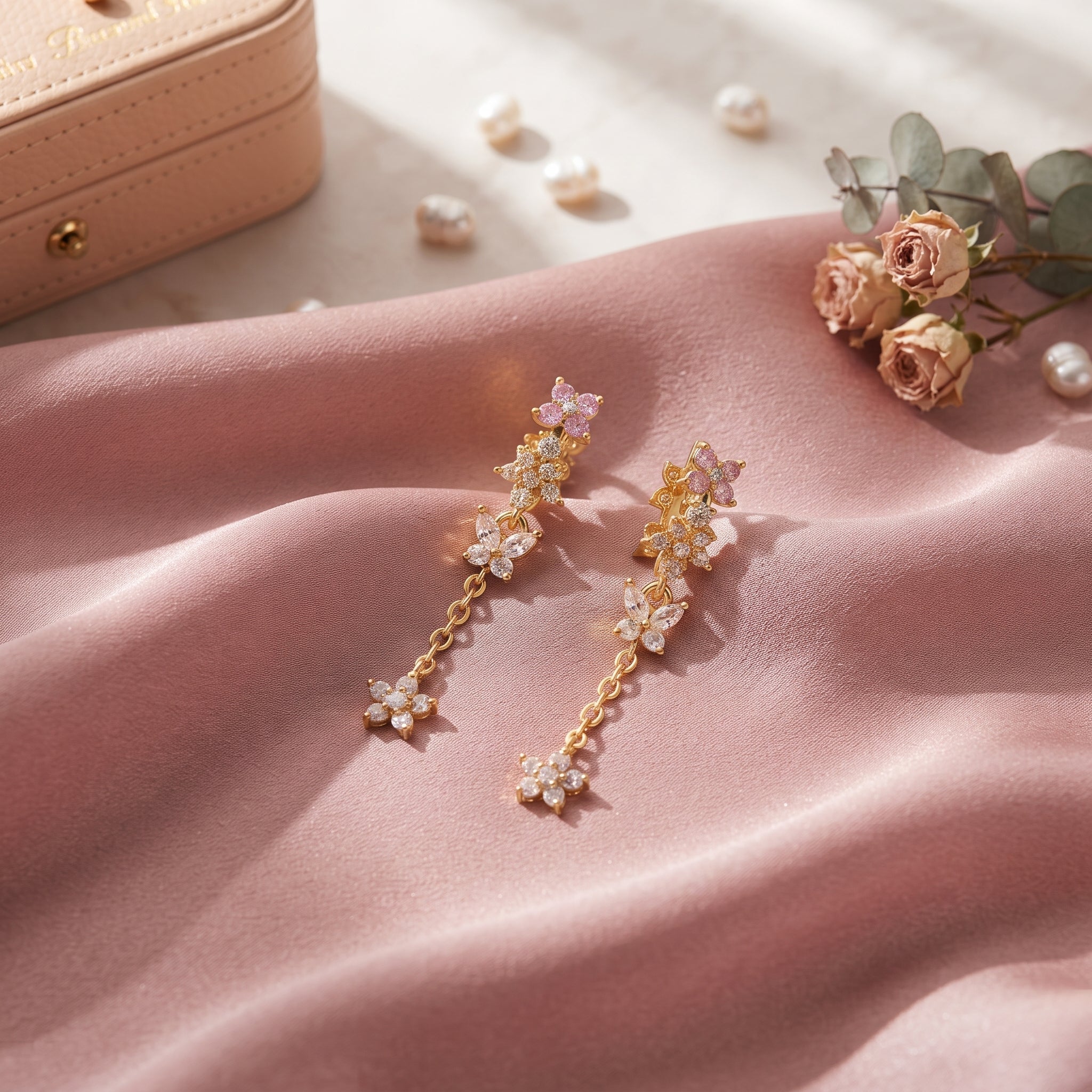A pair of gold dangle earrings with floral clusters and pink crystal-like accents, styled on draped pink fabric with scattered pearls, roses, and a beige jewelry case in the background.
