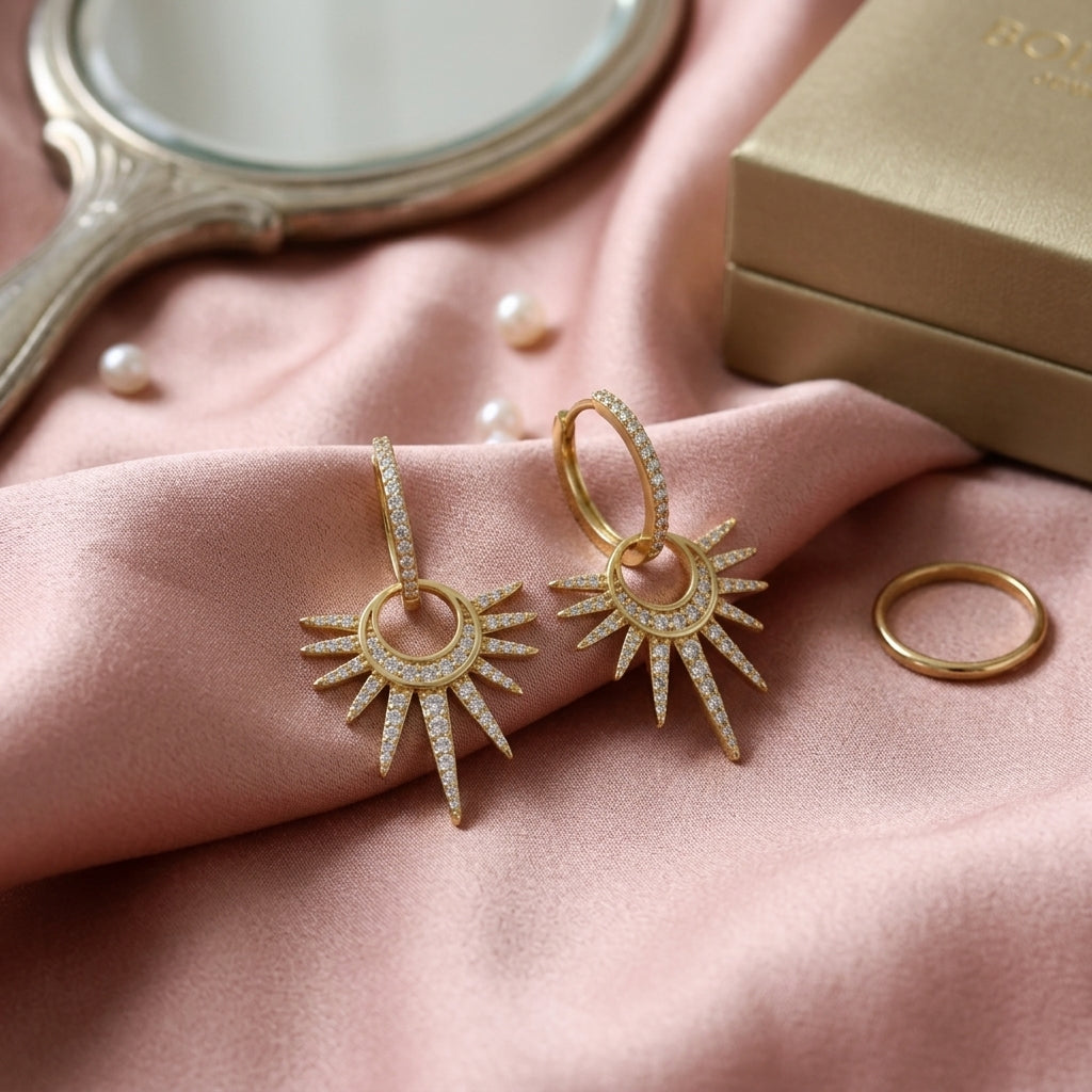 A pair of gold sunburst drop earrings with pavé-set clear stones, displayed on a draped pink satin fabric alongside a gold ring, a small mirror, pearls, and a gold jewelry box.