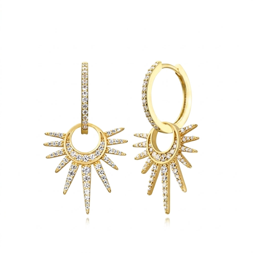 A pair of gold huggie hoop earrings with dangling sunburst charms, embellished with pavé-set clear cubic zirconia stones, shown on a solid white background.
