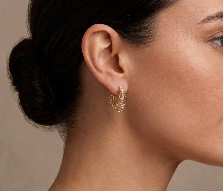 A close-up side profile of a woman with dark hair in a bun, wearing a delicate gold hoop earring with a textured, organic pattern, set against a neutral beige background.