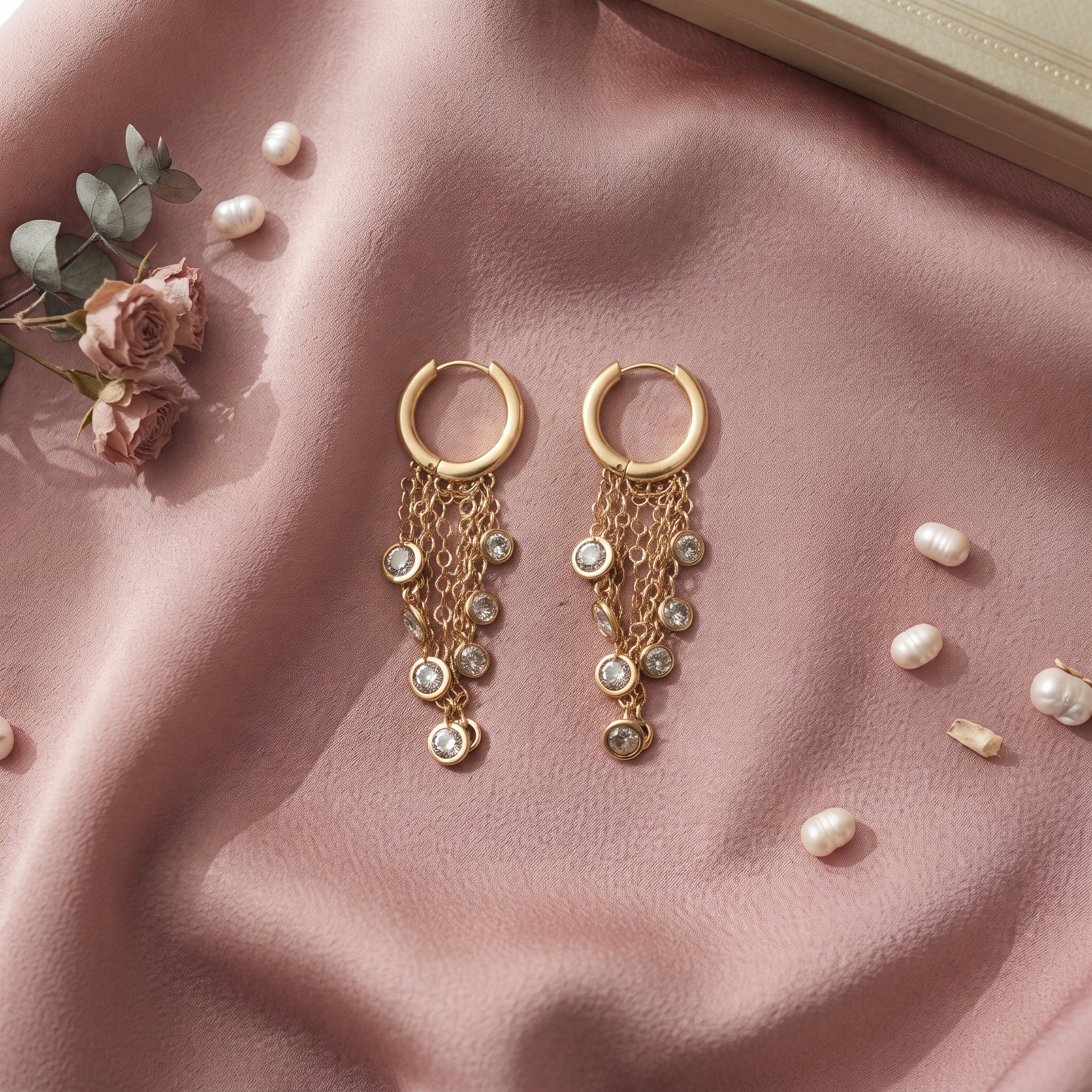 A pair of gold hoop dangle earrings with layered chain drops and small pearl-like beads, arranged on a pink satin fabric with scattered faux pearls, flowers, and a book corner in the background.