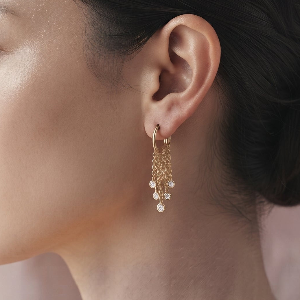 A woman in profile wearing a delicate gold dangle earring with small white pearl-like beads, set against a soft pink textured background.