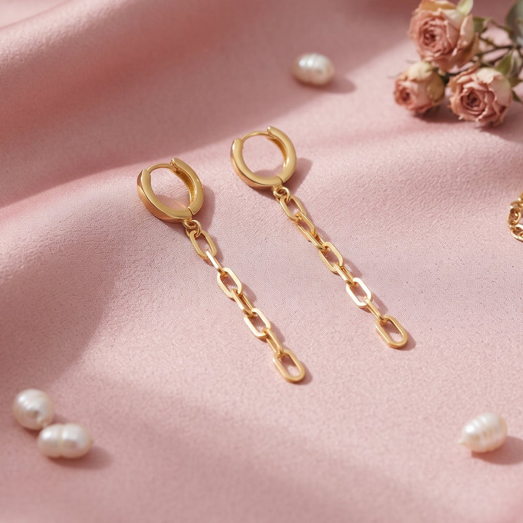 Gold hoop drop earrings with chain links, styled on a draped pink satin fabric with small pearls, roses, and a matching chain bracelet.