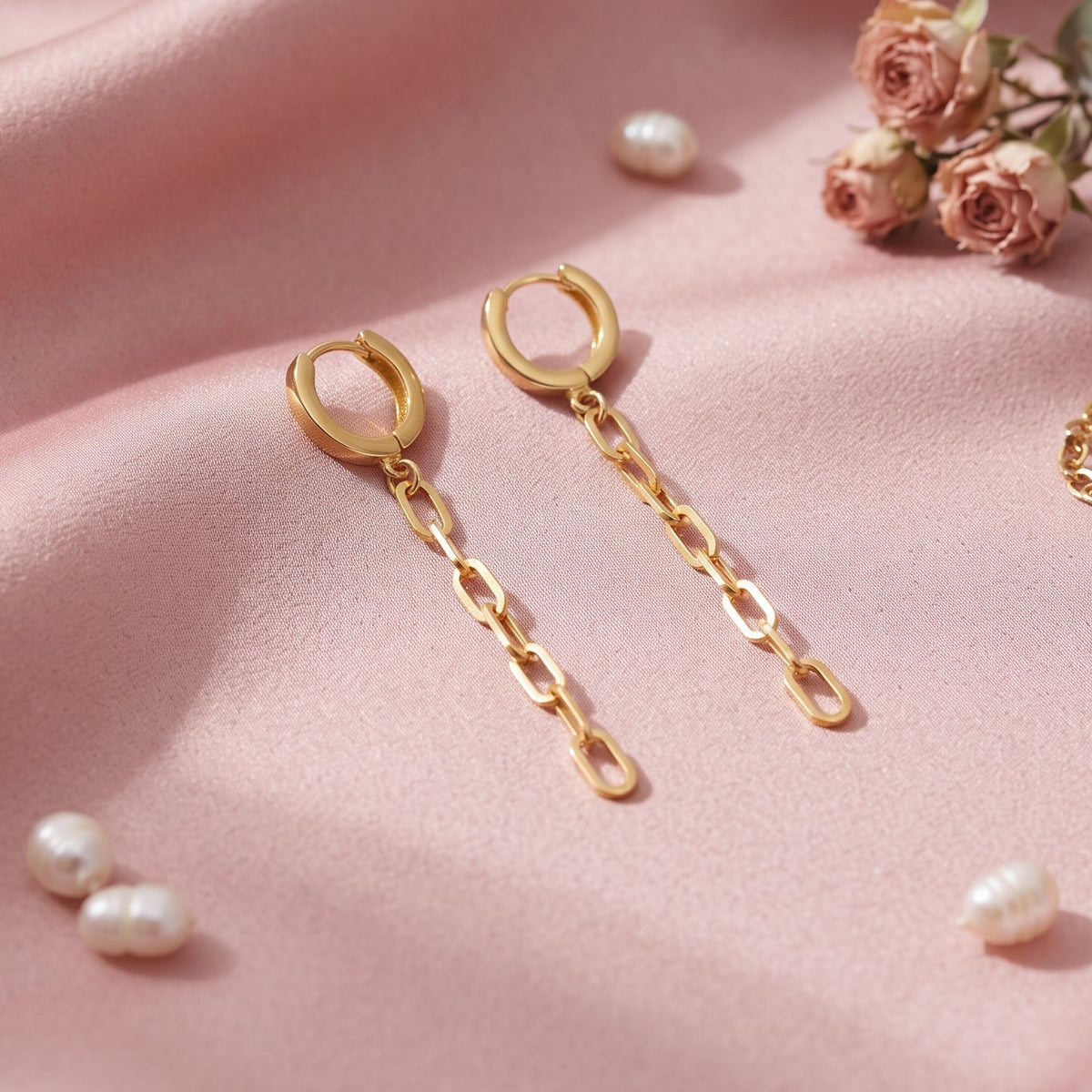 Gold hoop drop earrings with chain links, styled on a draped pink satin fabric with small pearls, roses, and a matching chain bracelet.