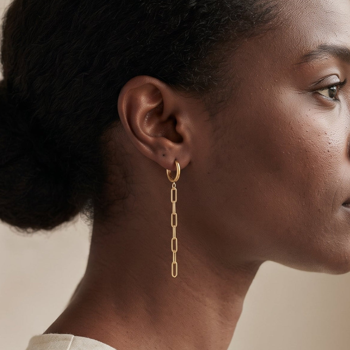 A woman in profile wearing a delicate gold dangle earring with a short chain link design, set against a soft beige background.