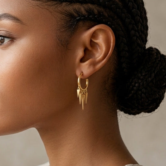 A woman in profile wearing a delicate gold hoop earring with three hanging teardrop charms, set against a neutral textured beige background.