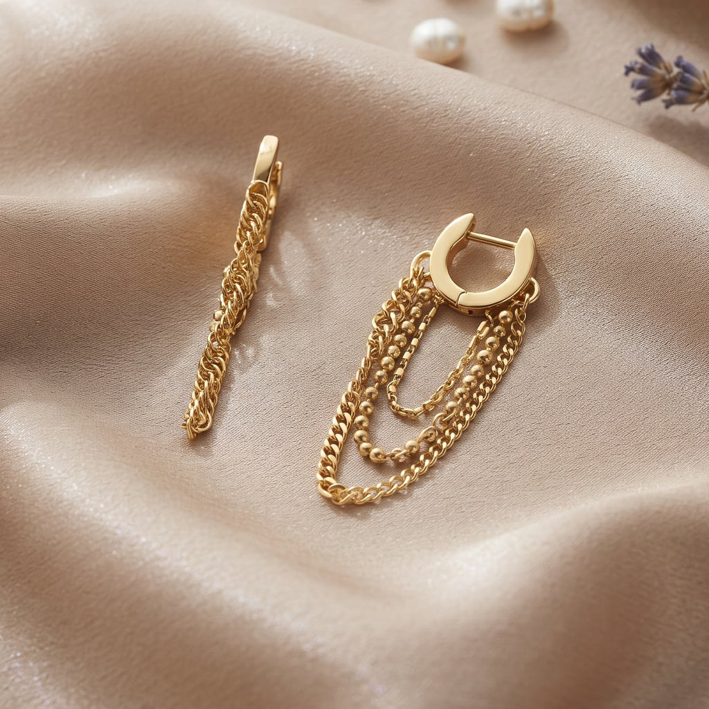 A pair of gold-toned huggie hoop earrings with layered chain and rope dangle details, displayed on draped champagne satin fabric with decorative pearls and dried lavender.