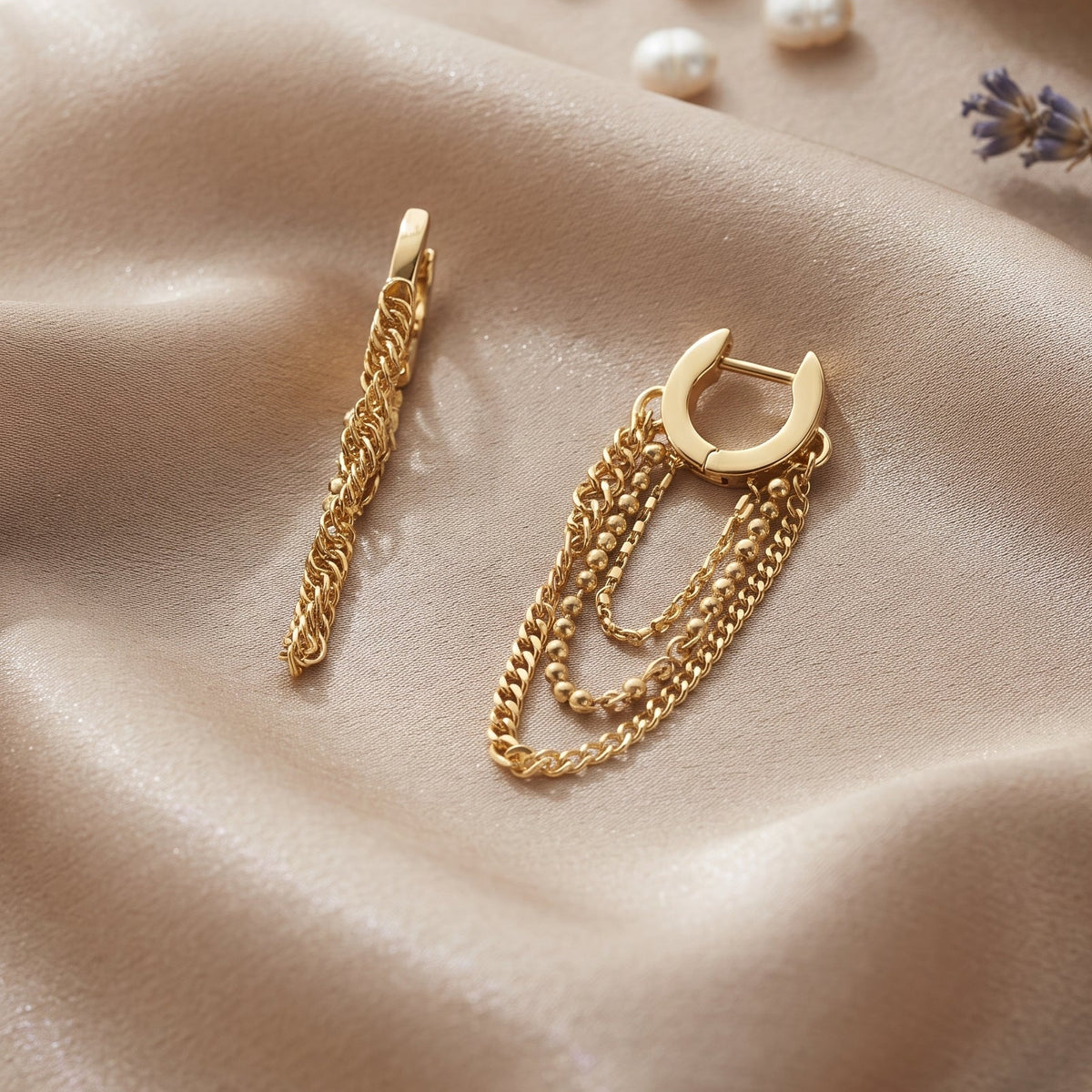 A pair of gold-toned huggie hoop earrings with layered chain and rope dangle details, displayed on draped champagne satin fabric with decorative pearls and dried lavender.