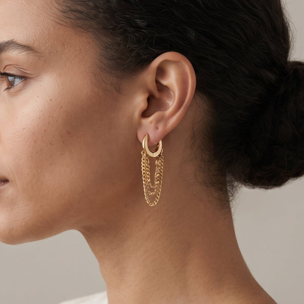 A woman in profile wearing a delicate gold huggie hoop earring with layered chain dangles, paired with a cream-colored linen top against a soft beige background.
