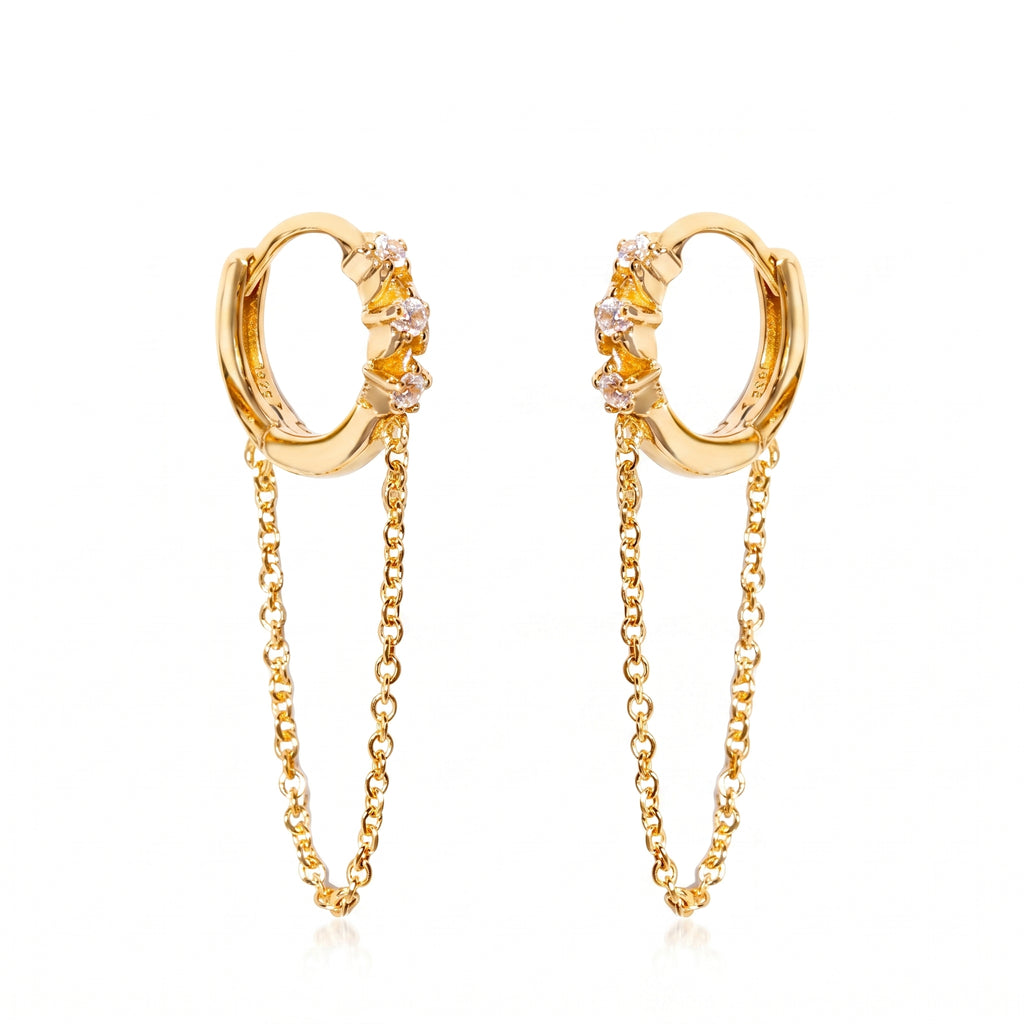 A pair of gold huggie hoop earrings with small clear gemstones and dangling chain accents, shown on a plain white background.