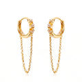 A pair of gold huggie hoop earrings with small clear gemstones and dangling chain accents, shown on a plain white background.