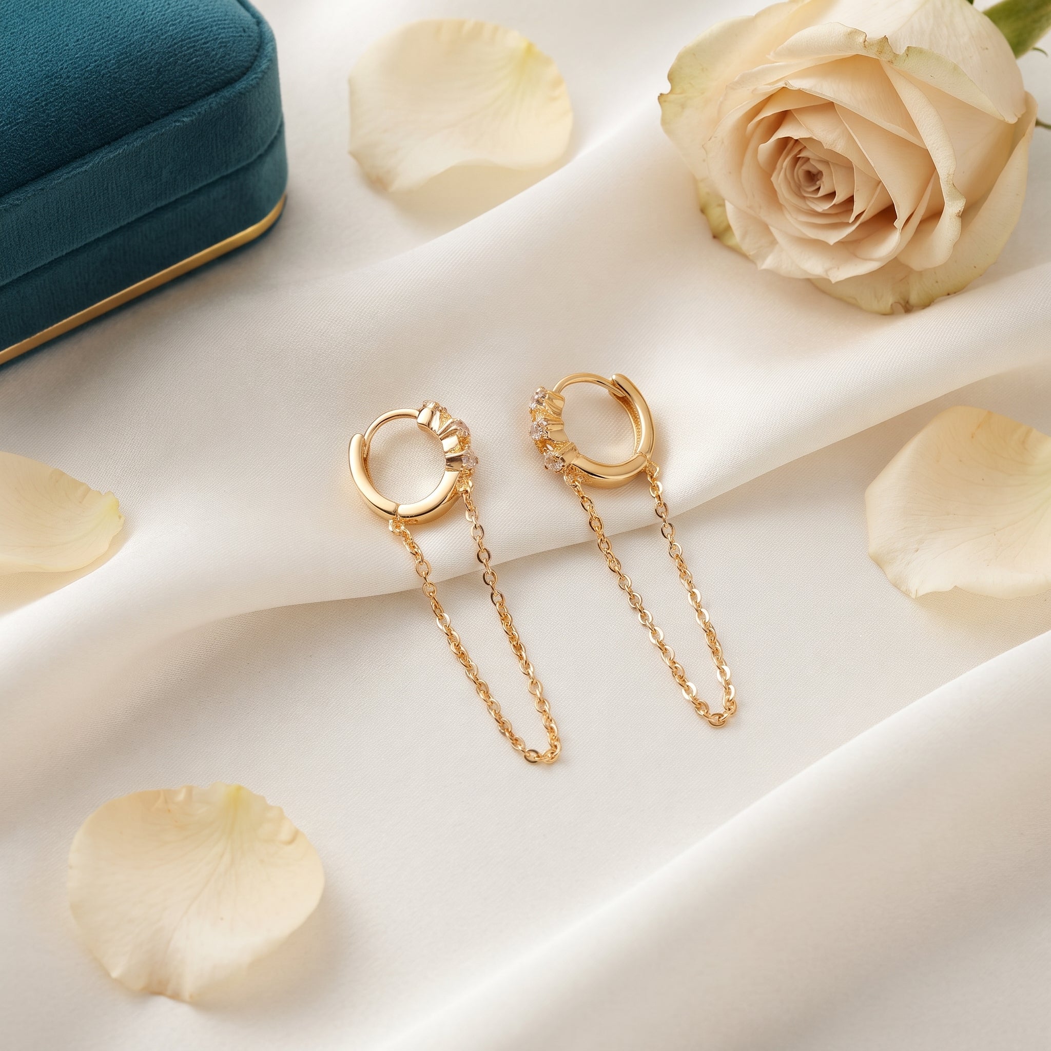 A pair of gold hoop earrings with delicate chain drops, styled on a draped white fabric background with cream rose petals, a cream rose, and a teal jewelry box.
